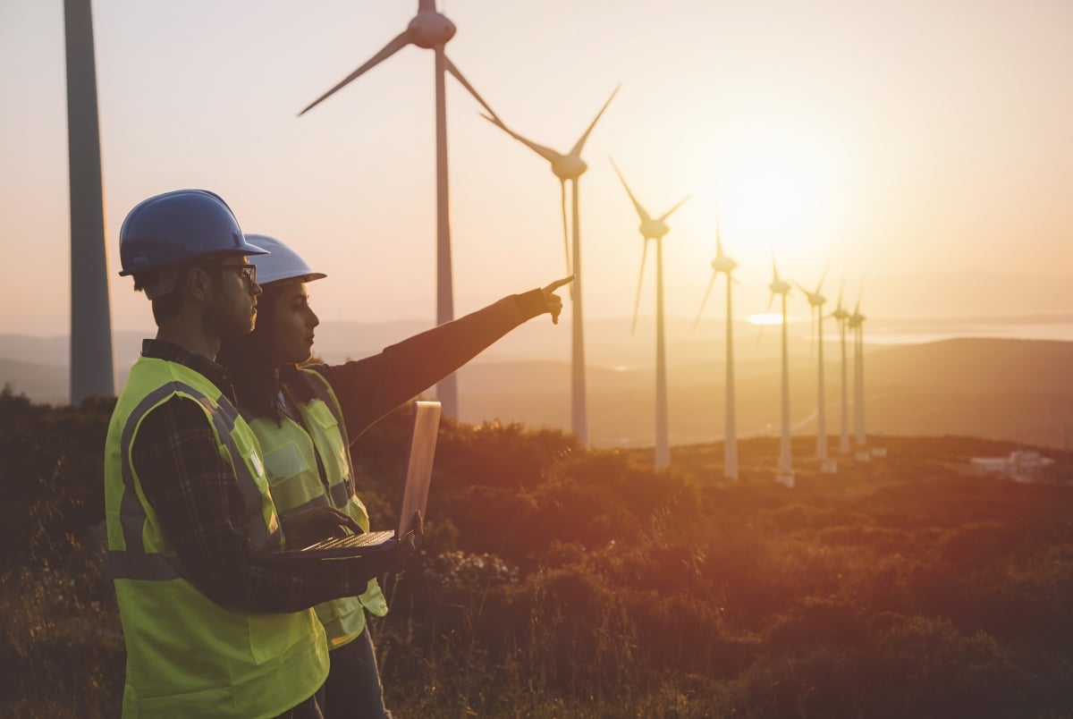 Two people, one pointing and one on a laptop, stand in front of a wind farm